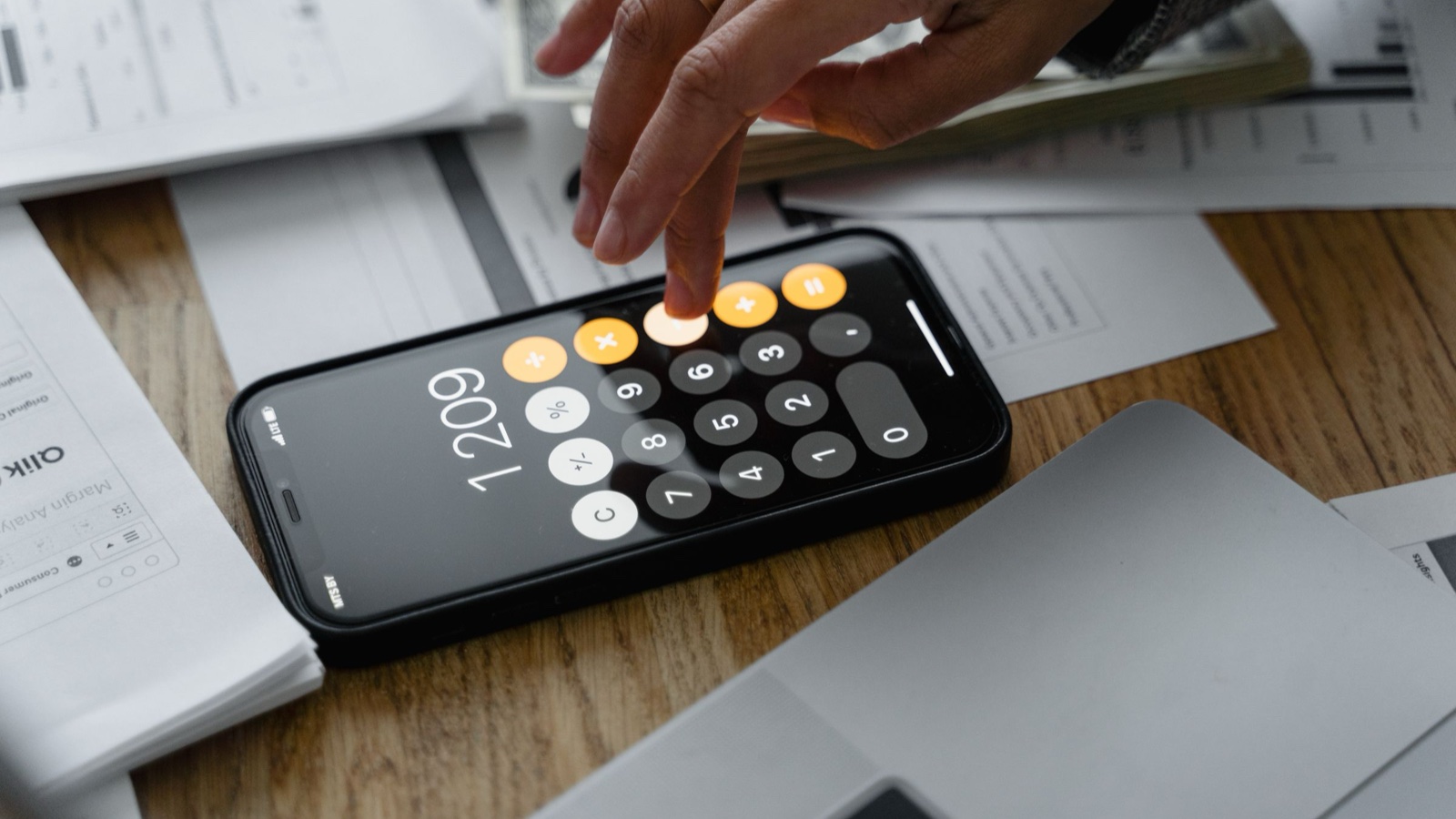 A finger taps the keys of a smartphone calculator lying on a wooden desk among printed papers.