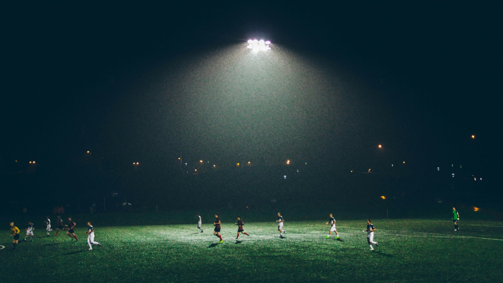 Players run across a floodlit pitch at night under a single bright floodlight.