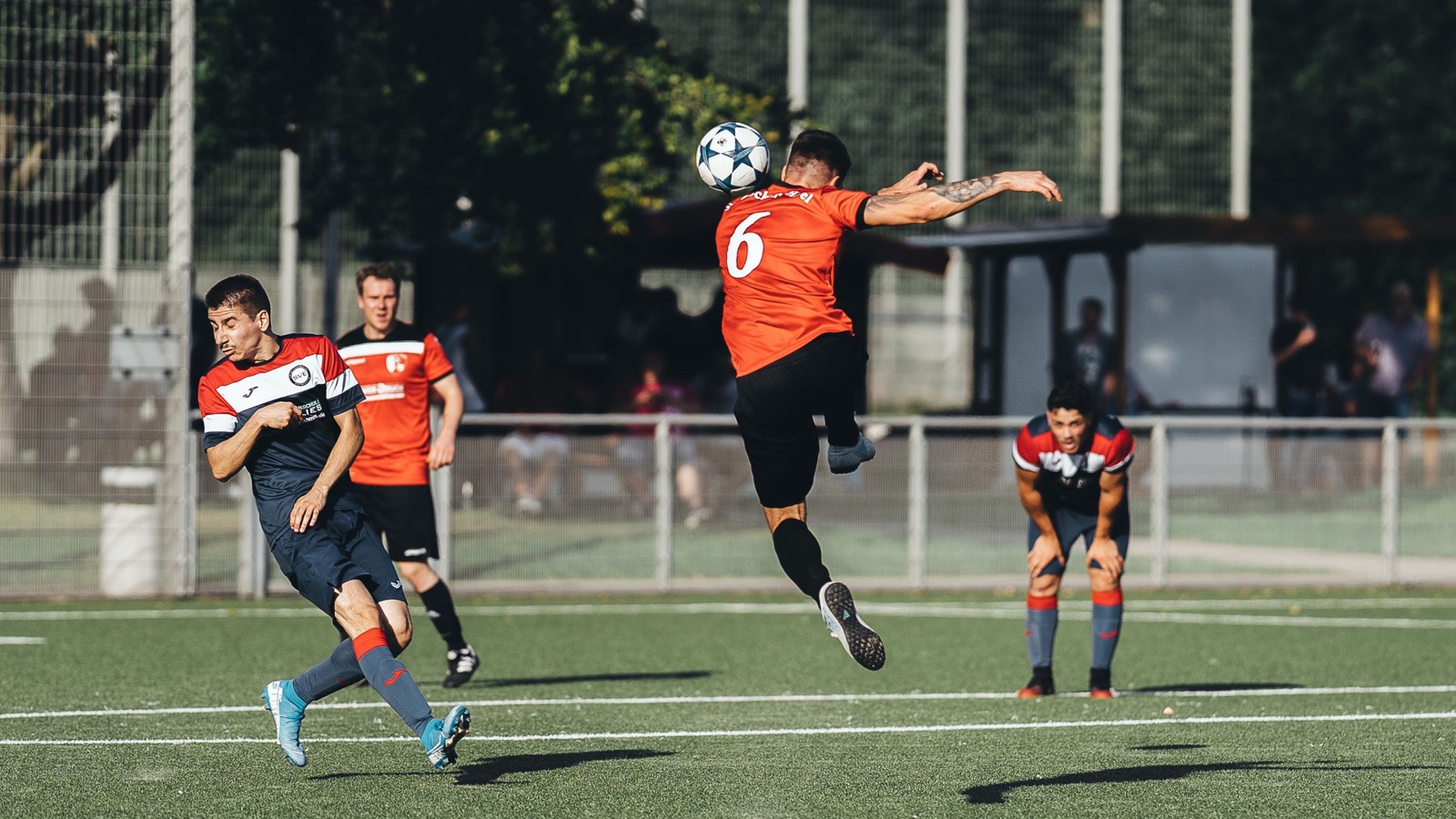 A footballer in an orange shirt leaps to head the ball while opponents watch.
