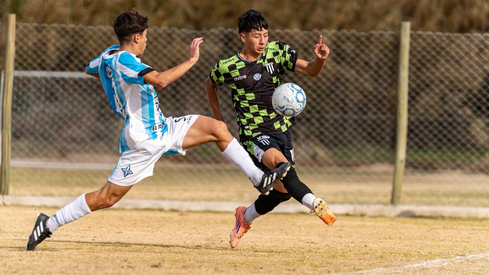 Two footballers in a tackle as one stretches a leg for the ball on a dry pitch.