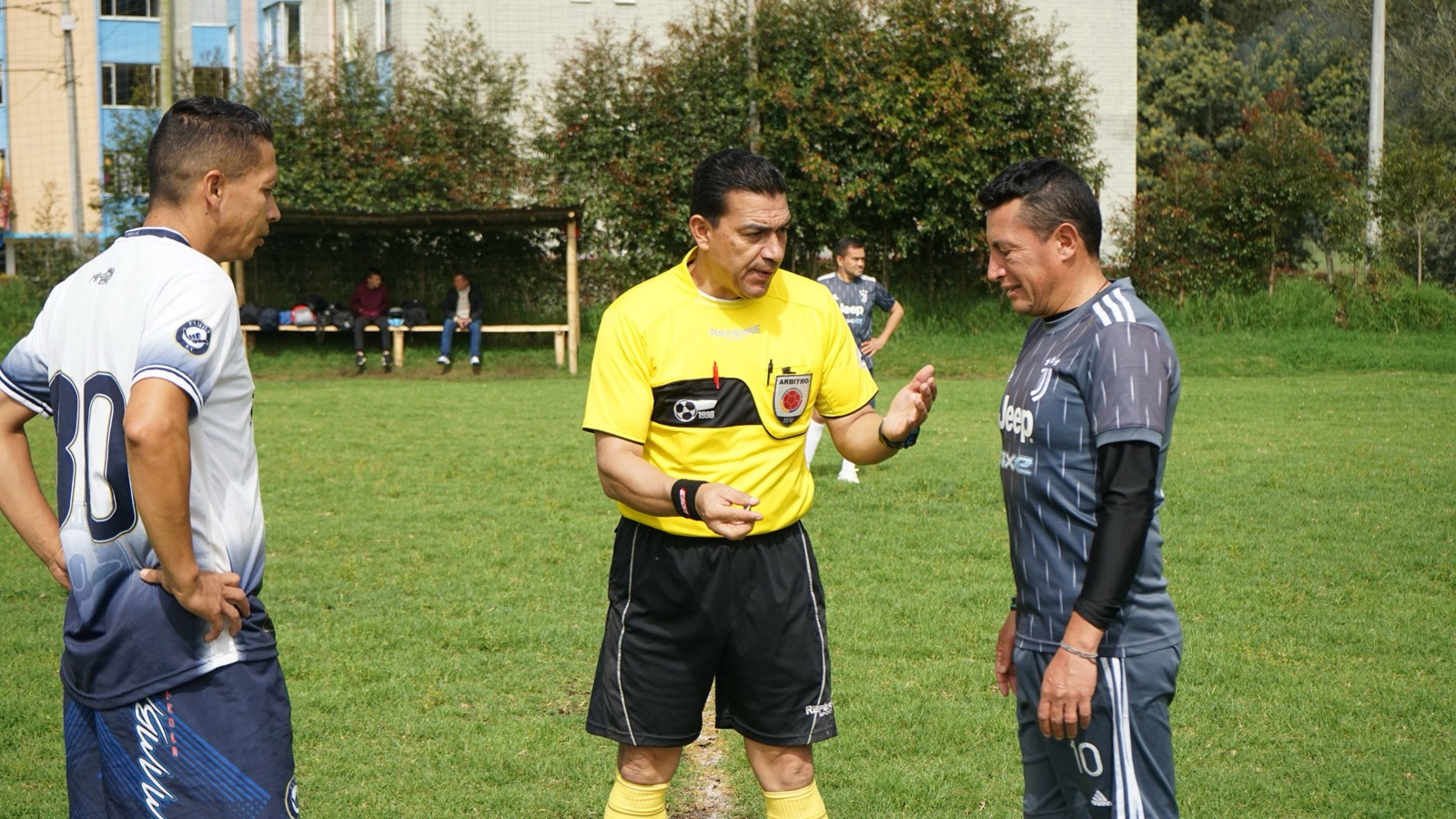 A referee in a yellow shirt speaks with two players on a grass pitch.