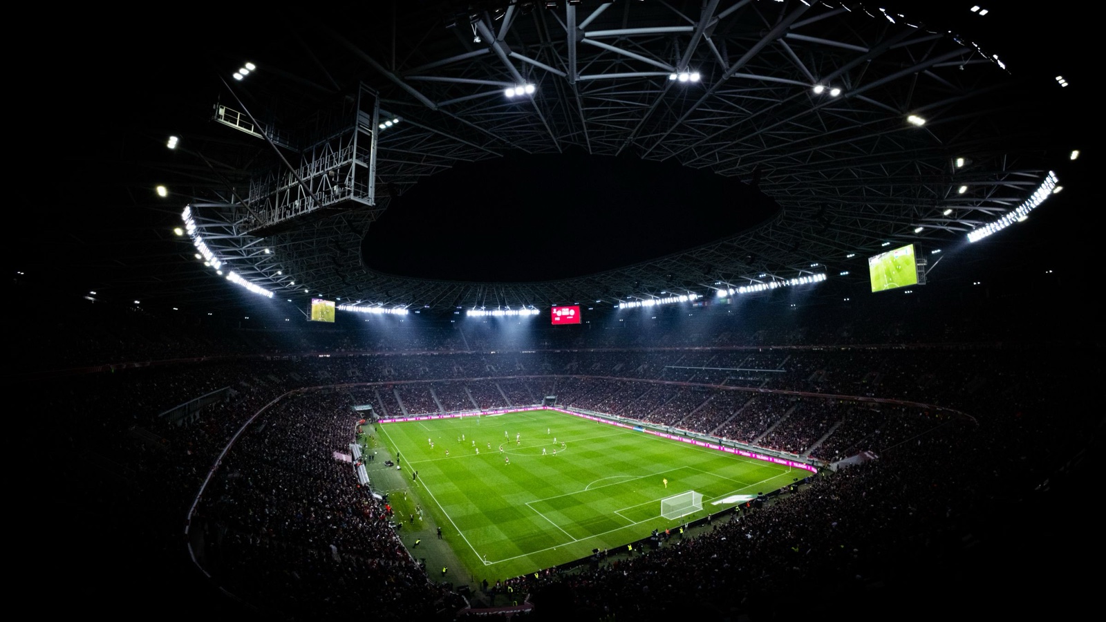 A floodlit football stadium at night with a packed crowd around the pitch.