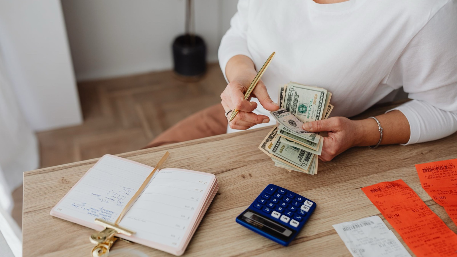 A woman holds a handful of dollar bills at a desk with a ledger, calculator and receipts.