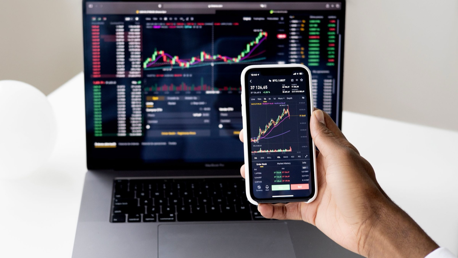 A hand holds a phone showing a crypto price chart in front of a laptop with another trading chart.