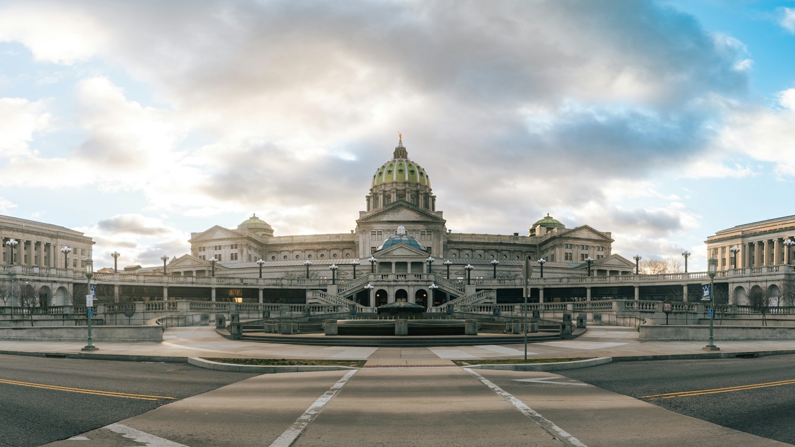 The domed Pennsylvania State Capitol in Harrisburg seen from the street under a cloudy sky.