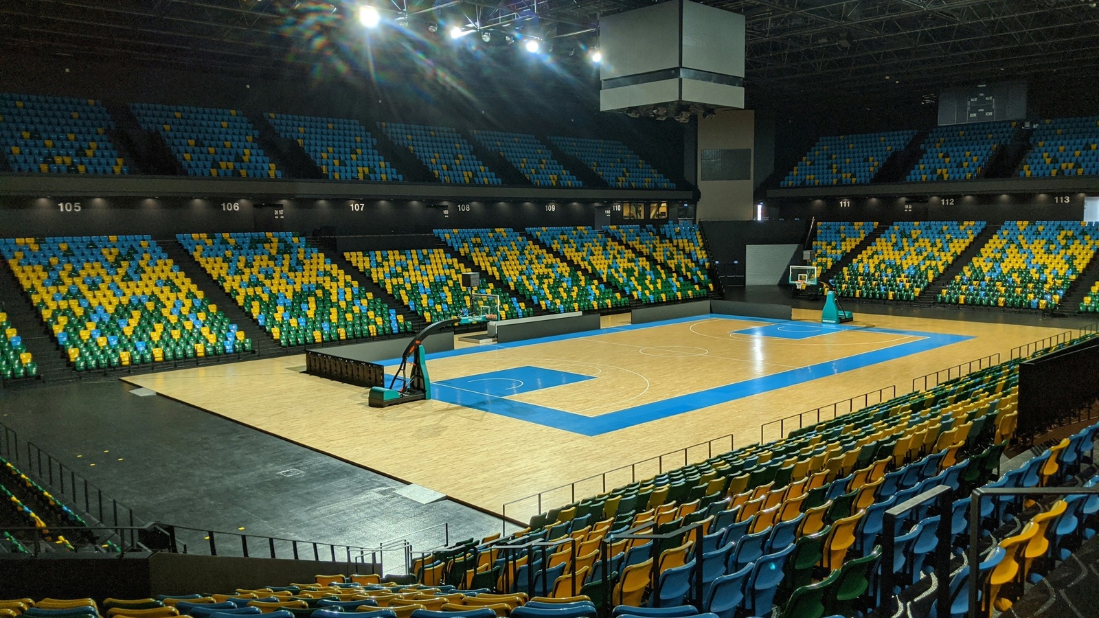 An empty indoor basketball arena with a blue-keyed wooden court and rows of blue, green and yellow seats under bright lights.