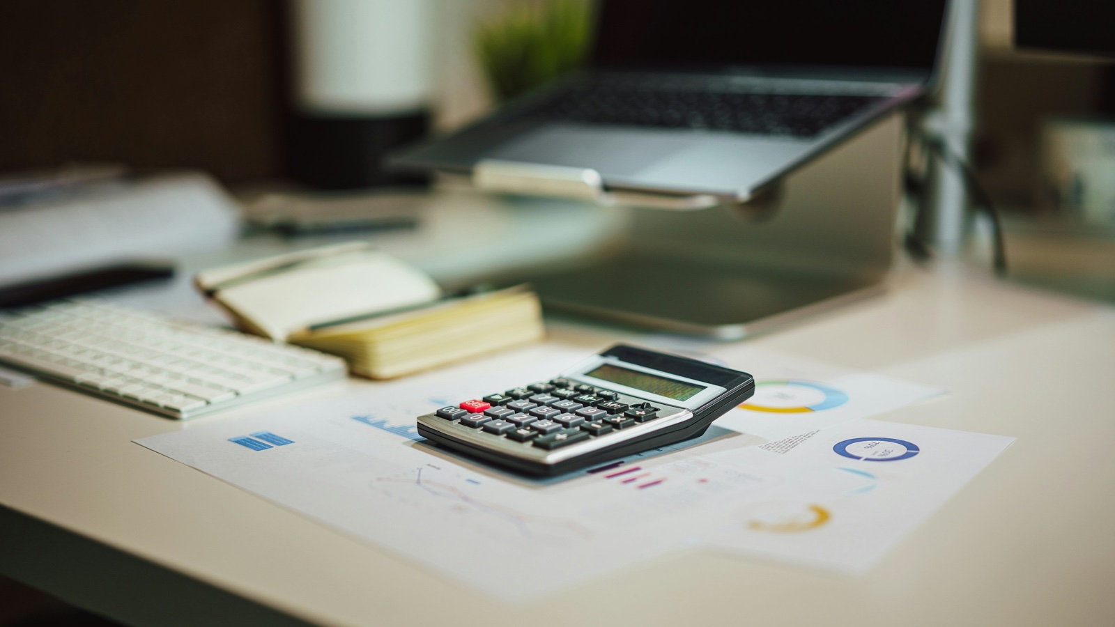 A pocket calculator resting on printed charts on a desk, with a laptop and notebook behind it.