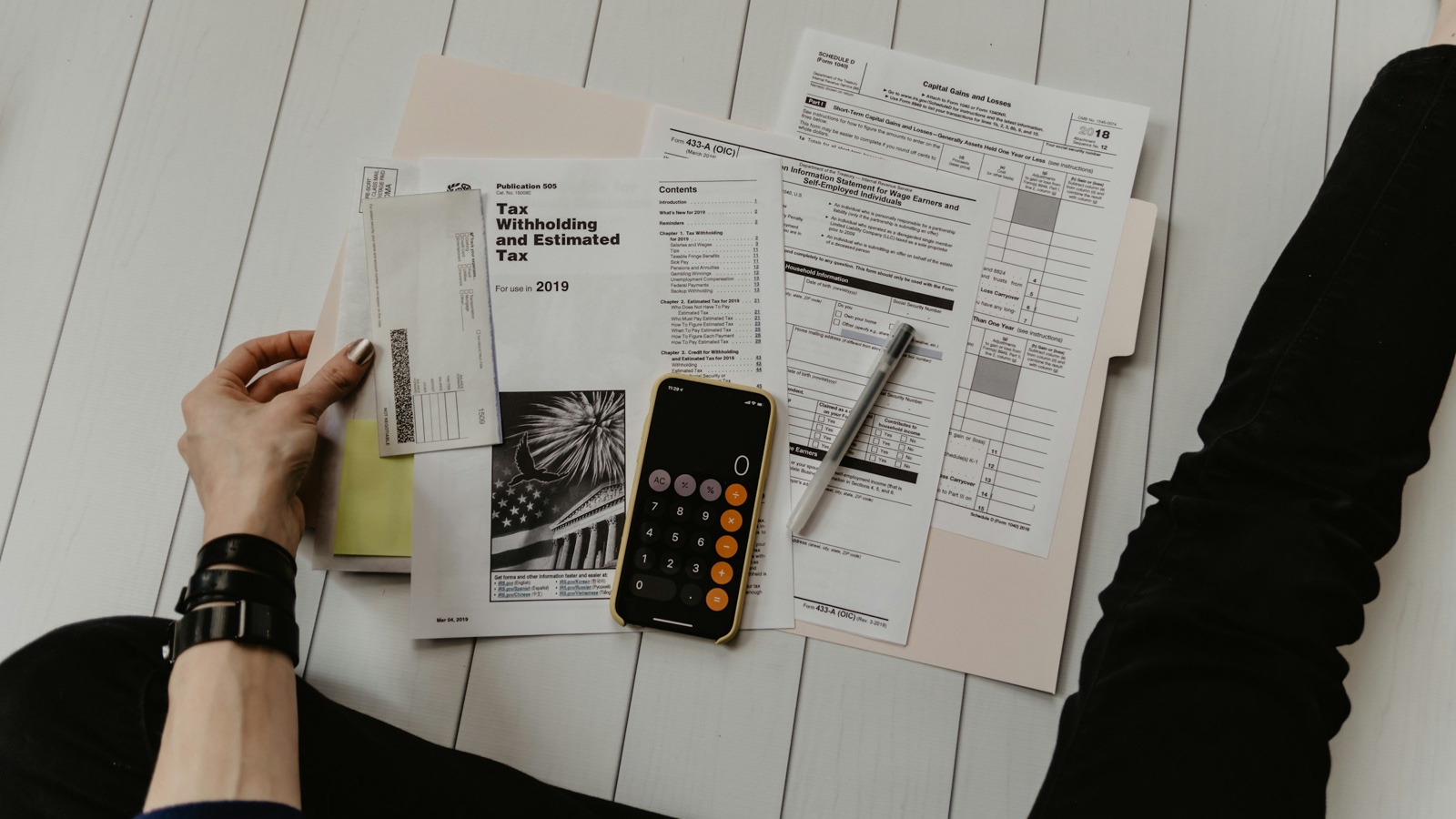 A hand holding an envelope beside US tax withholding forms, a pen and a phone showing a calculator on a white floor.