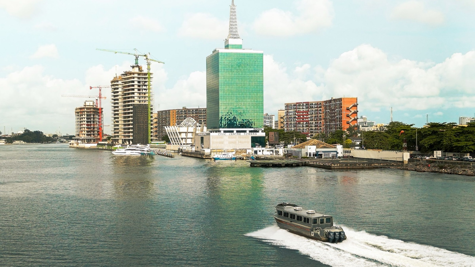 The Lagos waterfront with a glass office tower, cranes over new buildings and a boat crossing the lagoon.