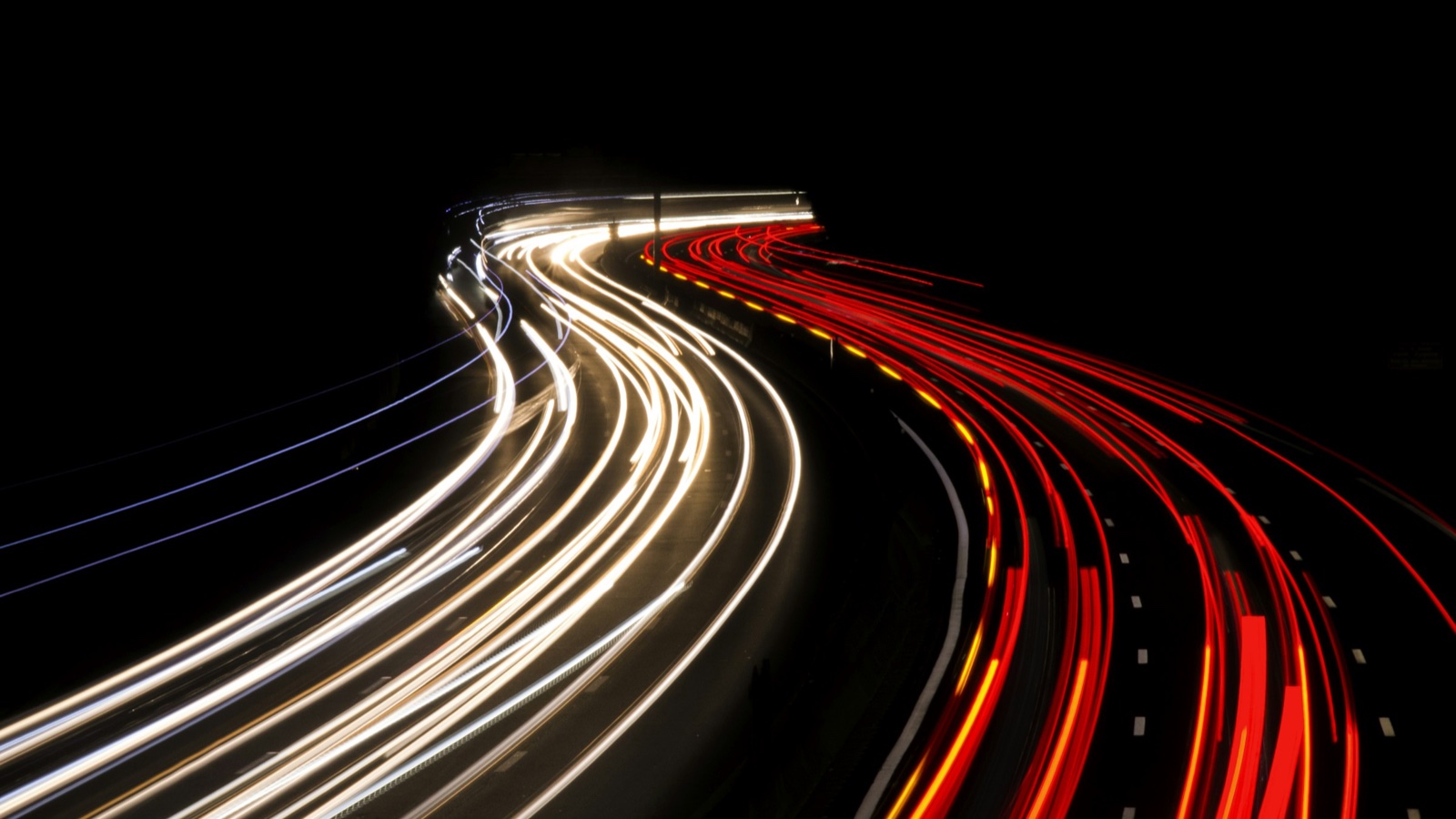 Long-exposure white and red light trails curve along a motorway at night.