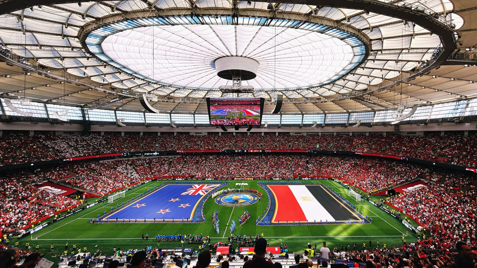 A packed stadium with a closed roof and giant New Zealand and Egypt flags spread across the pitch before a World Cup match.