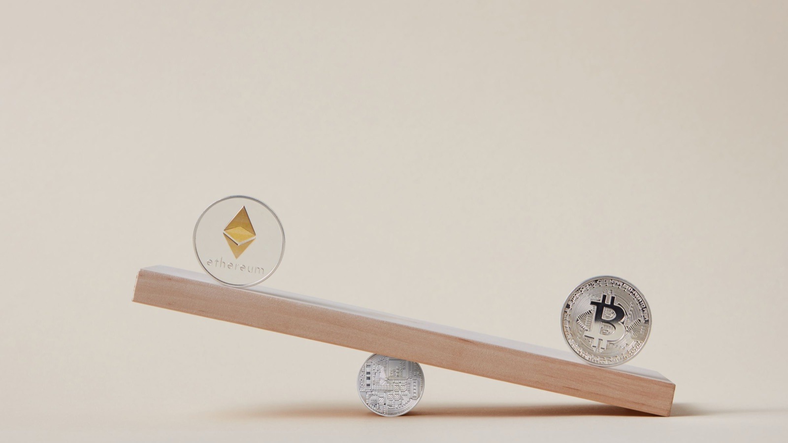 An Ethereum coin and a Bitcoin coin sit at opposite ends of a wooden plank tipped on a small coin.