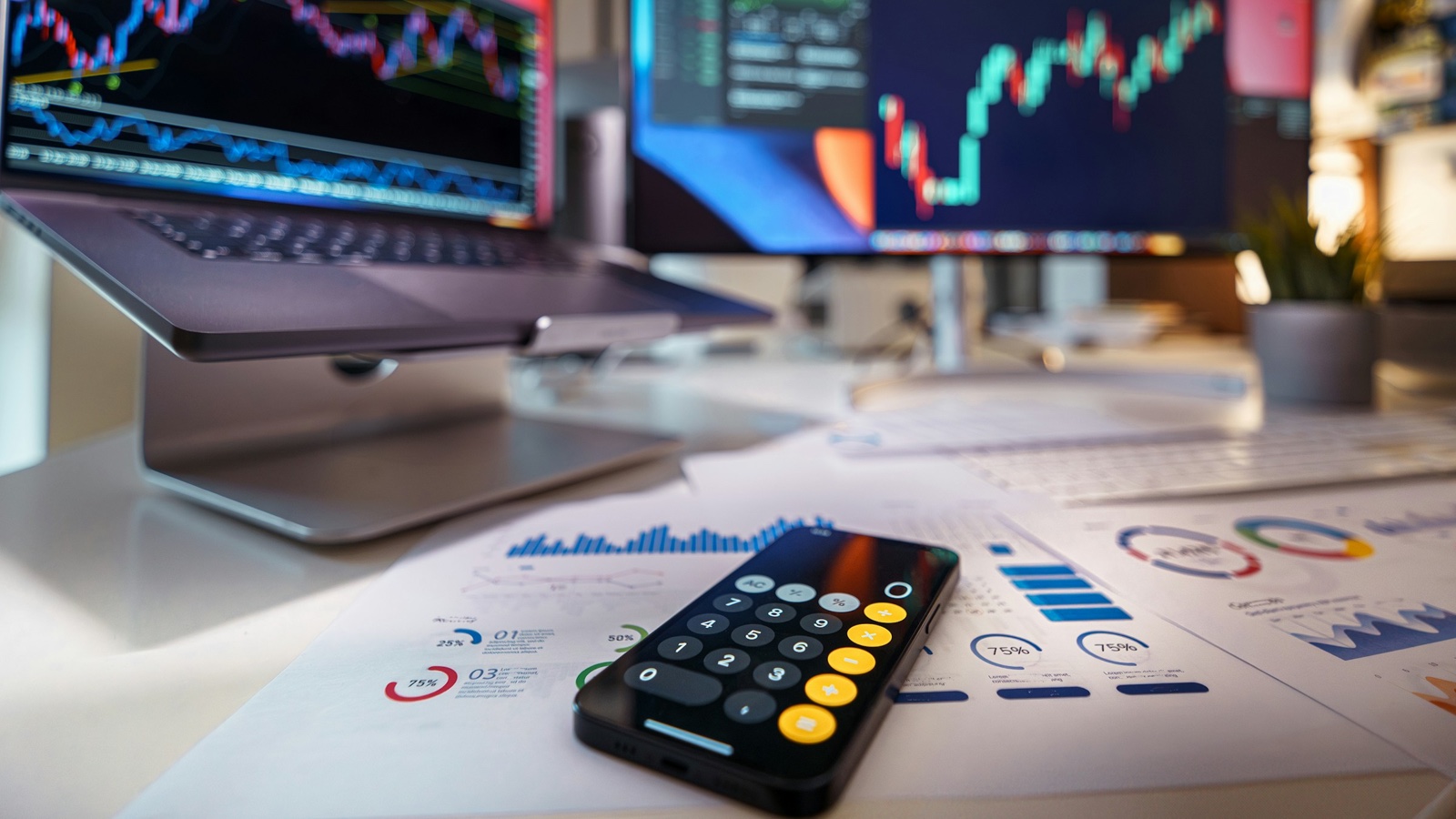 A smartphone calculator lies on printed charts on a desk in front of screens showing price charts.