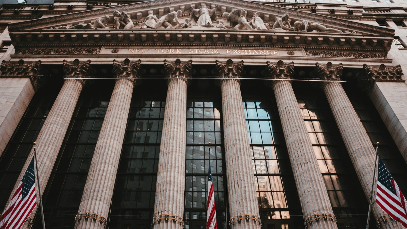 The columned front of the New York Stock Exchange seen from the street, with American flags at its base.