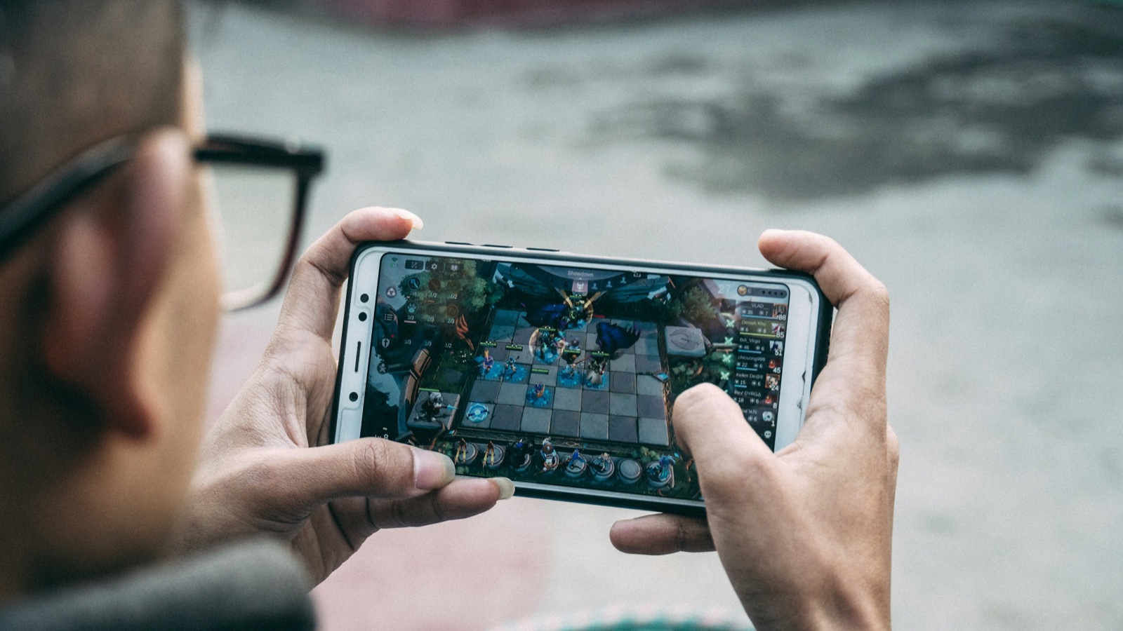 A man in glasses holds a phone sideways and plays a strategy game on its screen.