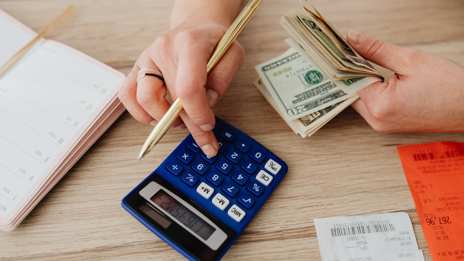 A person taps a calculator with a pen while holding a fan of twenty-dollar bills beside a notebook and receipts.