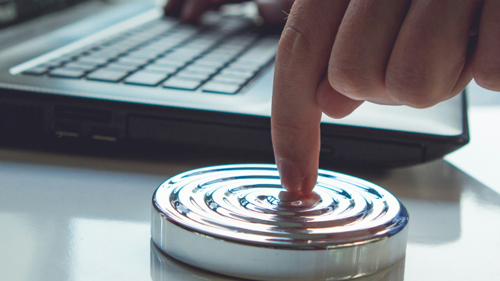 A finger pressing a large chrome button on a desk beside an open laptop