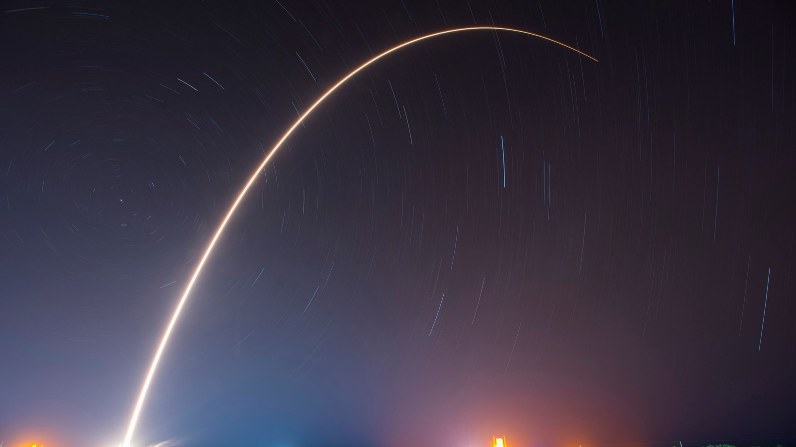 A long exposure of a rocket launch leaving a bright arc across a starry night sky.