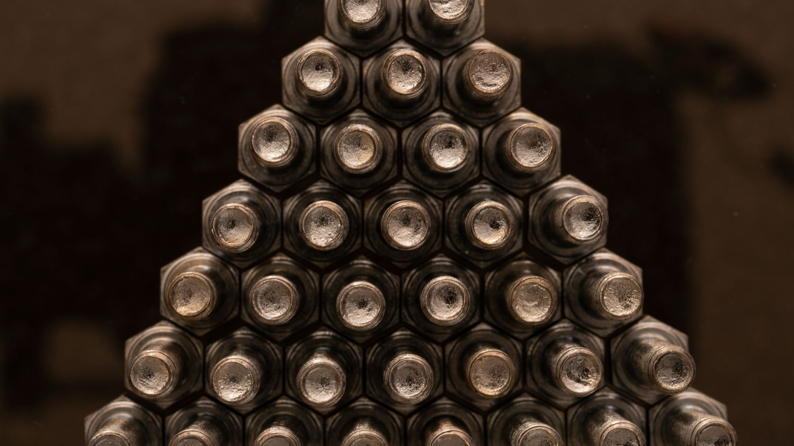 A triangle of metal bolts and nuts seen end on, arranged in rows that widen toward the bottom, like pegs on a drop board.