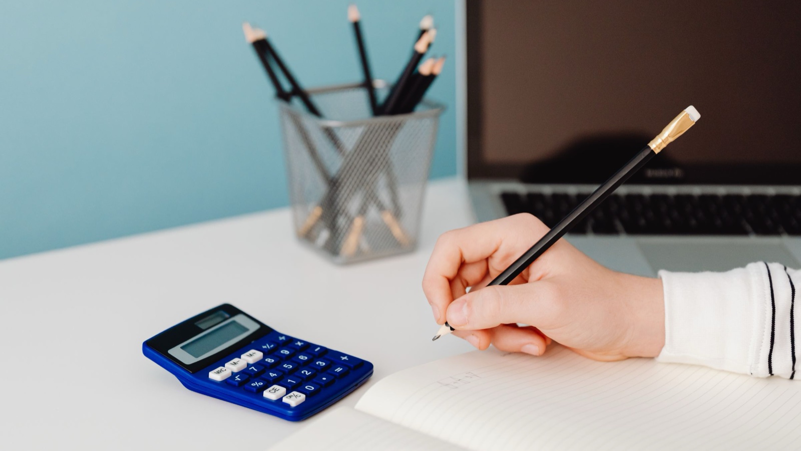 A hand writes with a pencil in a notebook beside a blue calculator and an open laptop on a white desk.