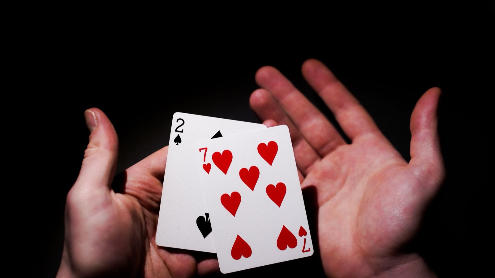Two open hands holding a two of spades and a seven of hearts against a dark background.