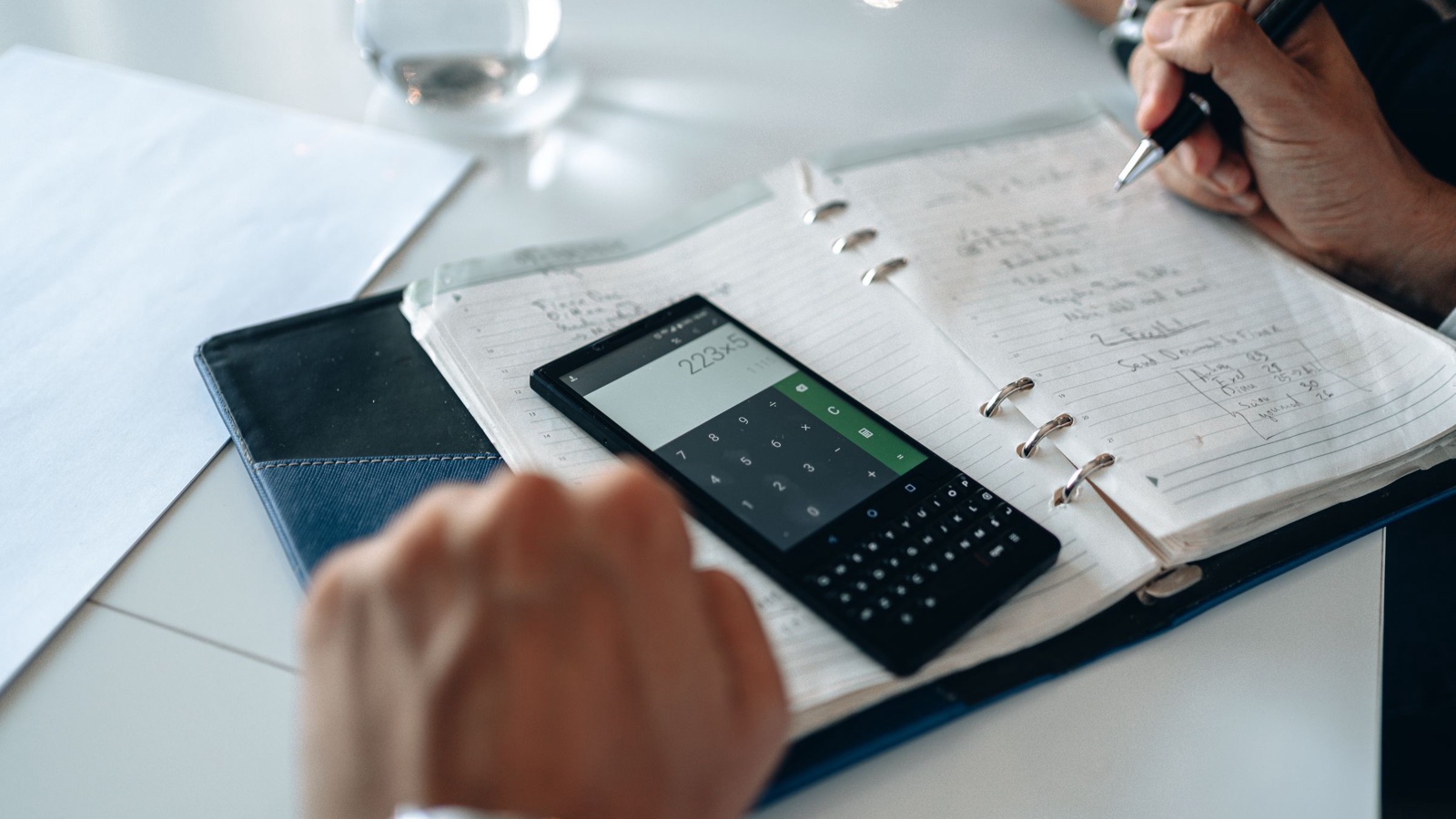A person writes figures in a notebook beside a phone showing a calculator.