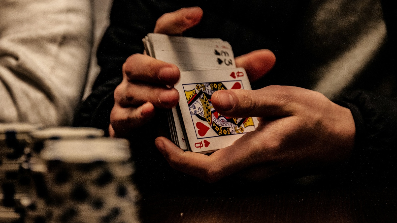 Two hands fan a short stack of playing cards, a queen of hearts showing, beside stacked chips at a dim table.