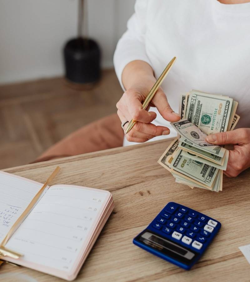 A woman holds a handful of dollar bills at a desk with a ledger, calculator and receipts.