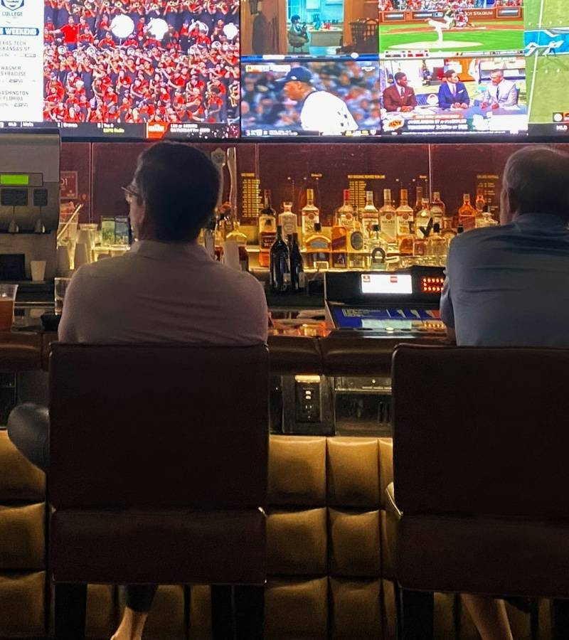 Four people sit at a sportsbook bar facing a wall of screens showing baseball and football games.