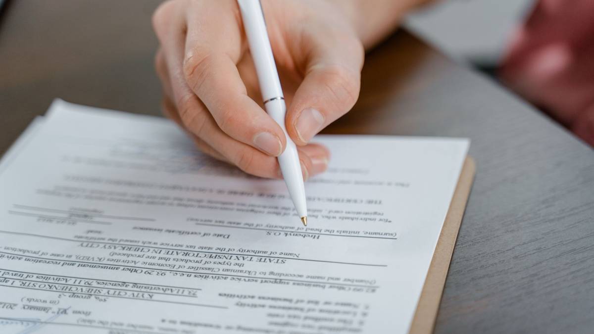 A hand holding a white pen over a printed form on a clipboard, ready to sign.