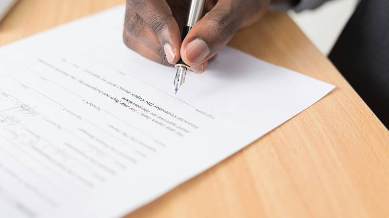 A hand with a fountain pen about to sign a form with several signature lines on a wooden desk.