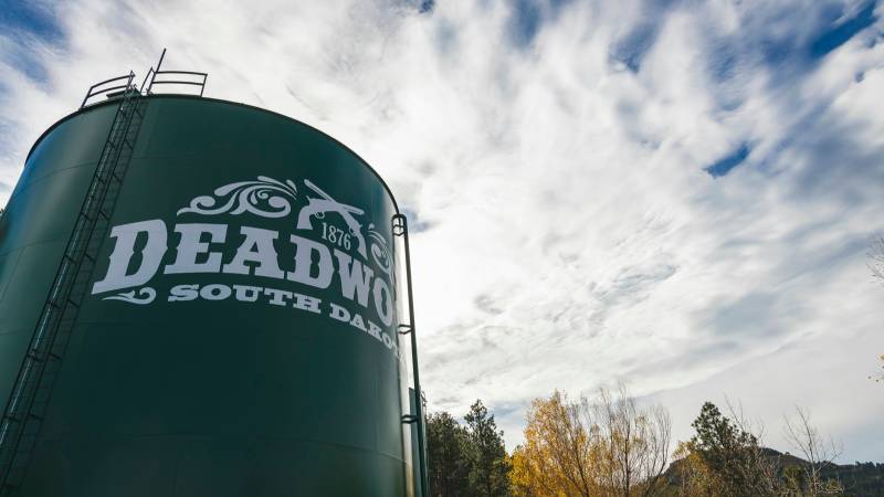 A green water tower painted with the Deadwood, South Dakota 1876 logo under a cloudy blue sky.