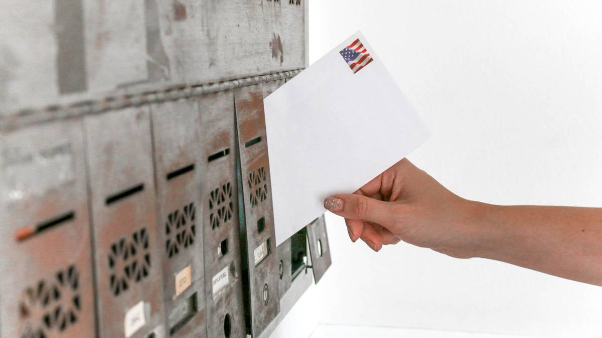 A hand slides a stamped envelope into a row of metal mailboxes.