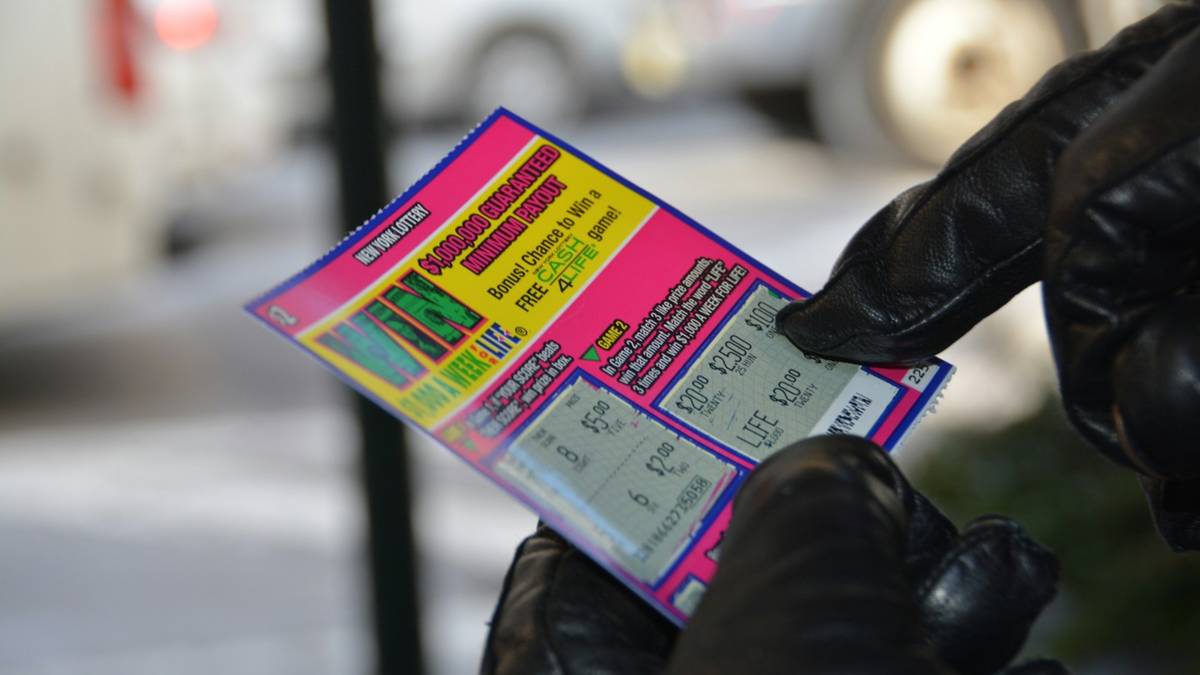 A gloved hand holds a pink New York Lottery scratch-off ticket with its panels partly scratched.