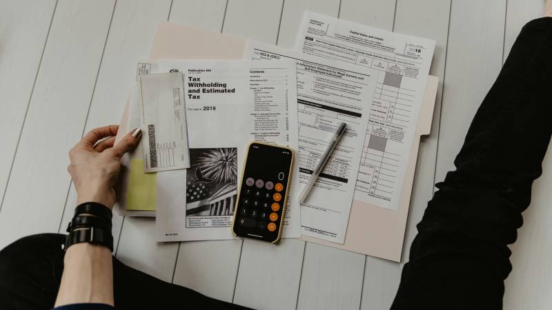 A hand holding an envelope beside US tax withholding forms, a pen and a phone showing a calculator on a white floor.