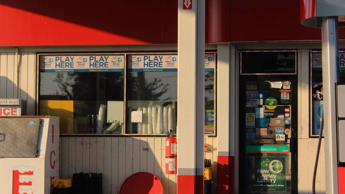 A gas station storefront in New Jersey with Play Here lottery posters in the window and a New Jersey Lottery sticker on the door