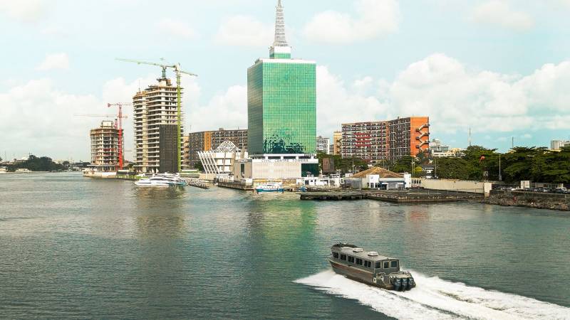 The Lagos waterfront with a glass office tower, cranes over new buildings and a boat crossing the lagoon.