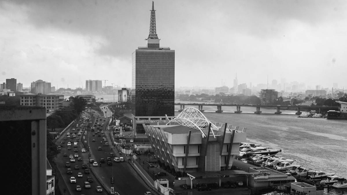 Black and white view of Lagos with a tall tower, a busy road and the lagoon.