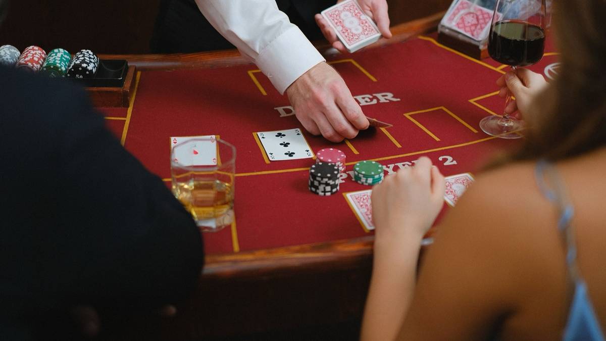 A dealer deals cards at a red felt casino table with chips and drinks.