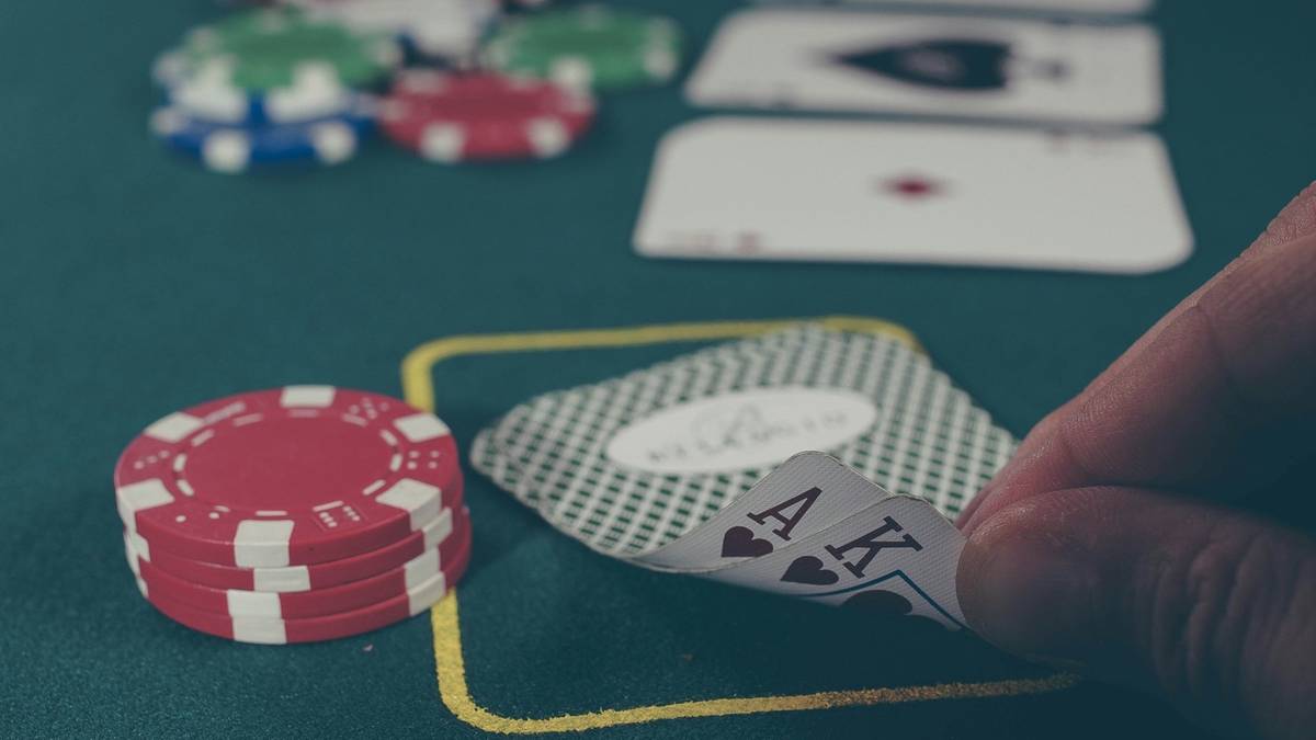 A hand lifts the corner of an ace and king of hearts beside a stack of red chips on a green poker table.
