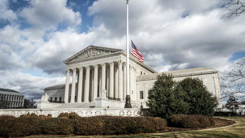 The white marble columns of the US Supreme Court building stand under a cloudy sky beside a flagpole.
