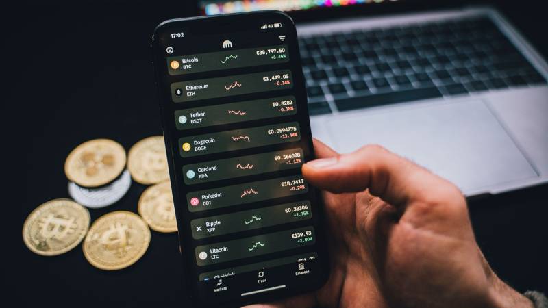 A hand holds a phone showing a list of cryptocurrency prices, with gold coins and a laptop on a dark desk.