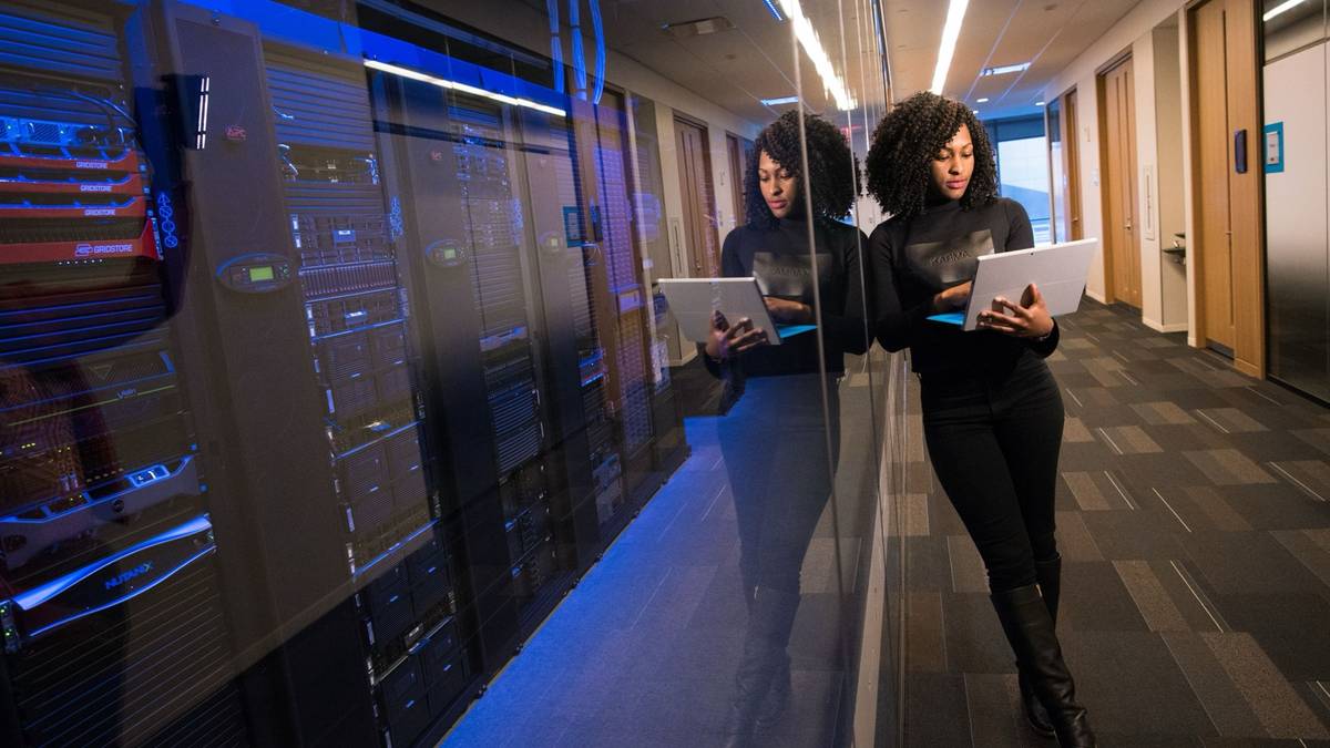 A woman stands in a glass-walled data centre corridor reading from a laptop.