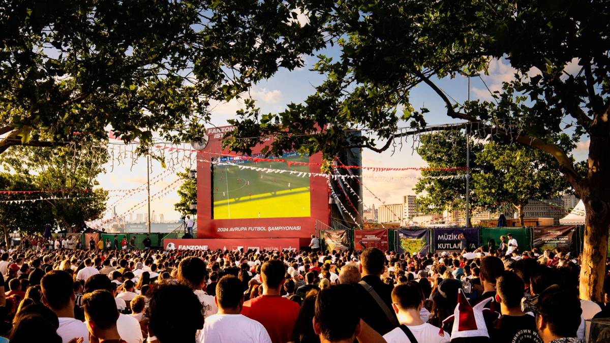 A large crowd sits under trees in a park watching a football match on a big outdoor screen.