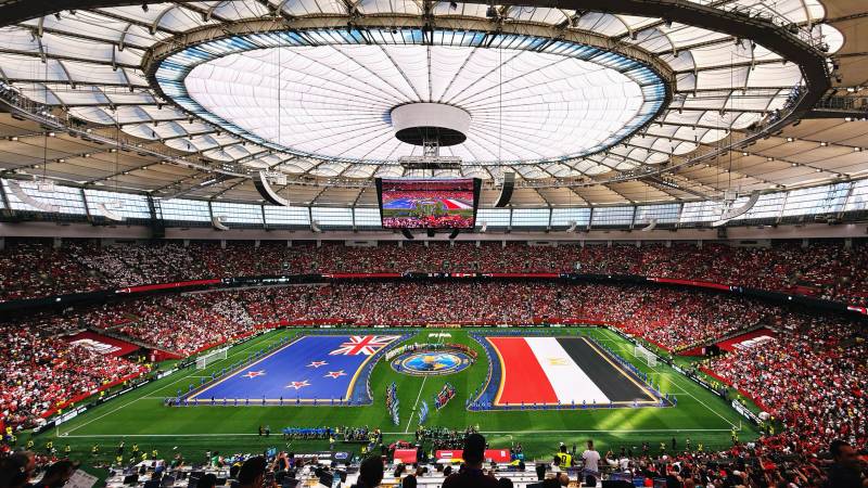 A packed stadium with a closed roof and giant New Zealand and Egypt flags spread across the pitch before a World Cup match.