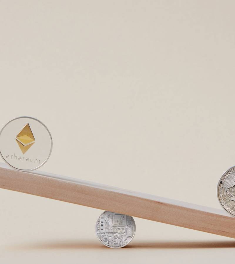 An Ethereum coin and a Bitcoin coin sit at opposite ends of a wooden plank tipped on a small coin.