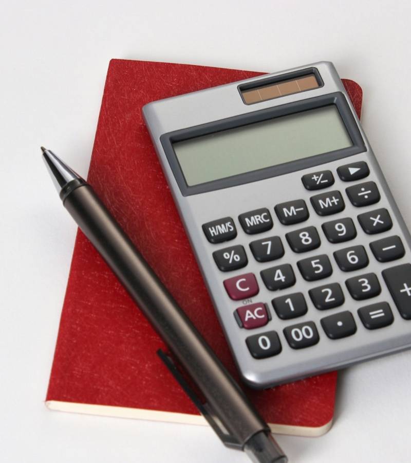 A silver pocket calculator and a black pen resting on a red notebook on a white desk.