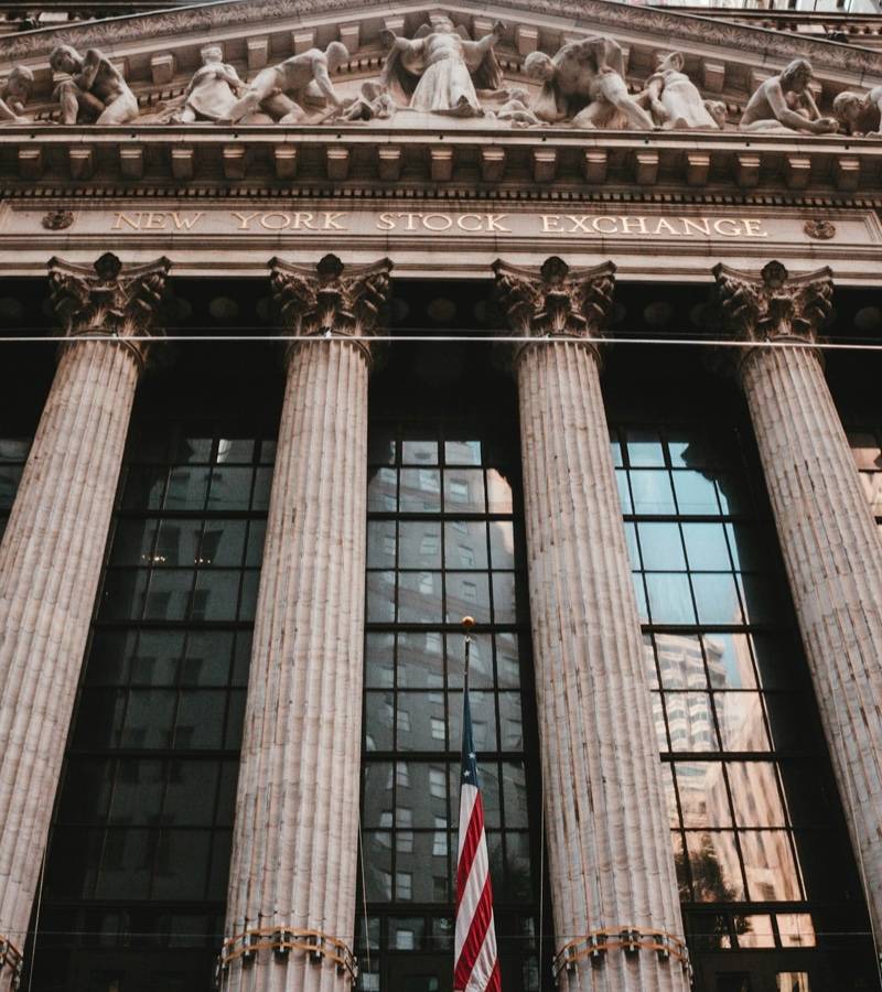 The columned front of the New York Stock Exchange seen from the street, with American flags at its base.