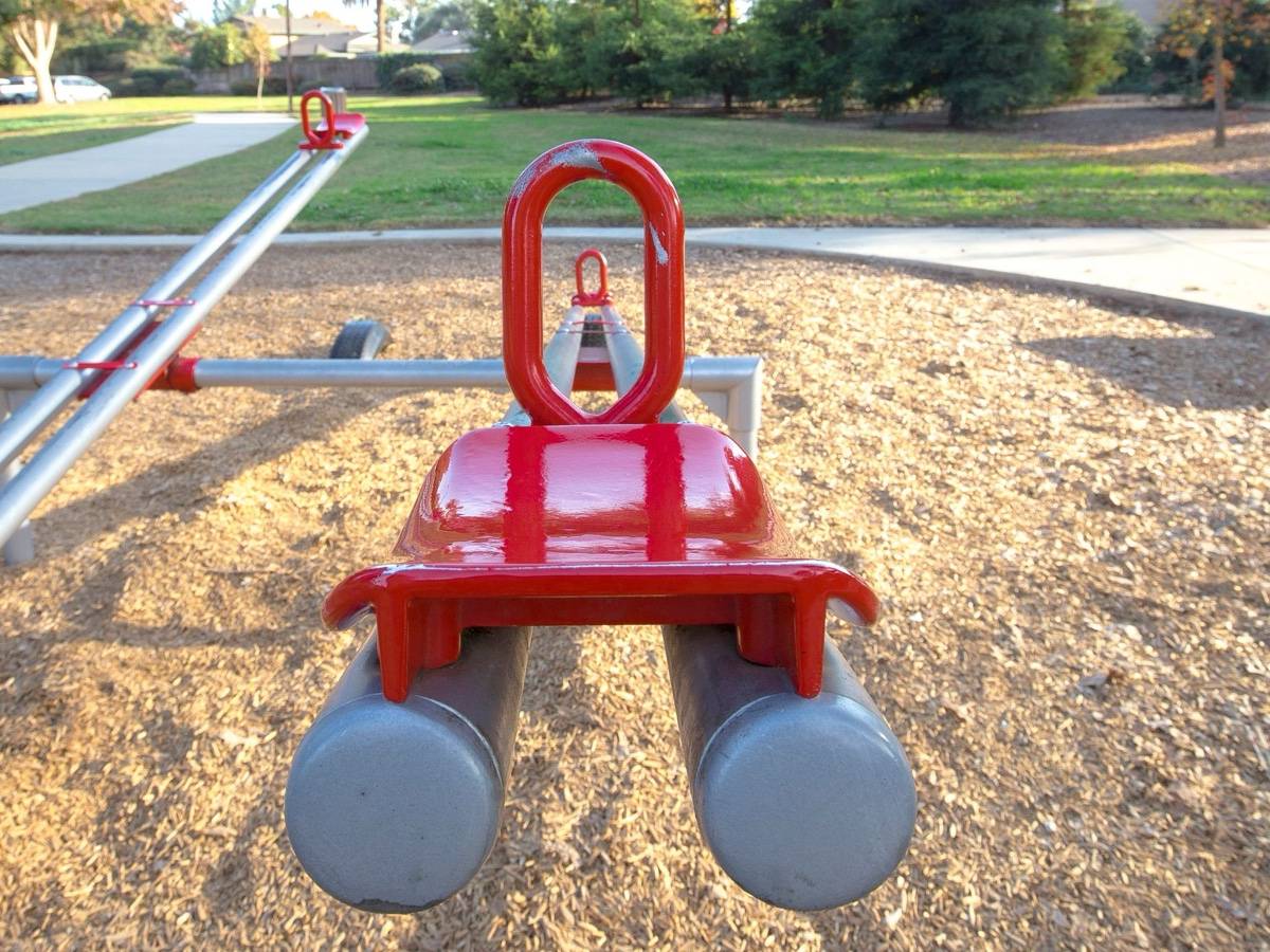 A red seesaw seat seen close up on a playground with wood chip ground.