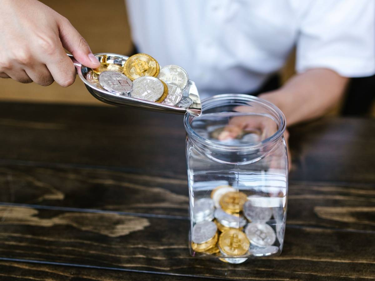 A hand tips a scoop of gold and silver coins into a glass jar on a dark wooden table.